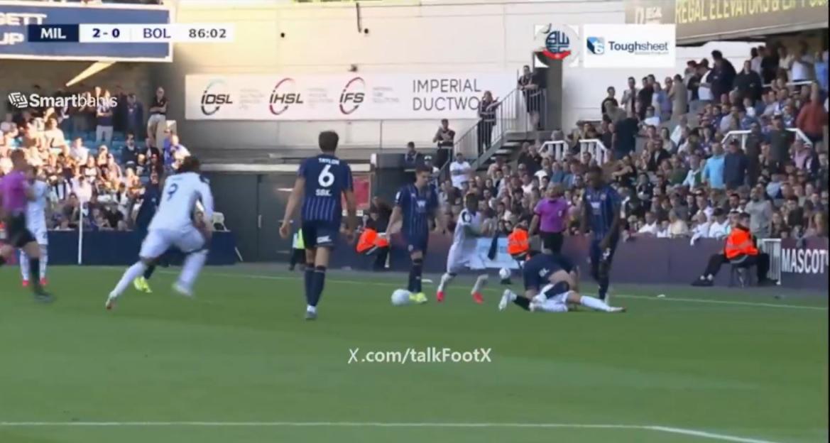 A soccer player in a white uniform falls to the ground in front of a player in a blue uniform, while other players and fans look on.