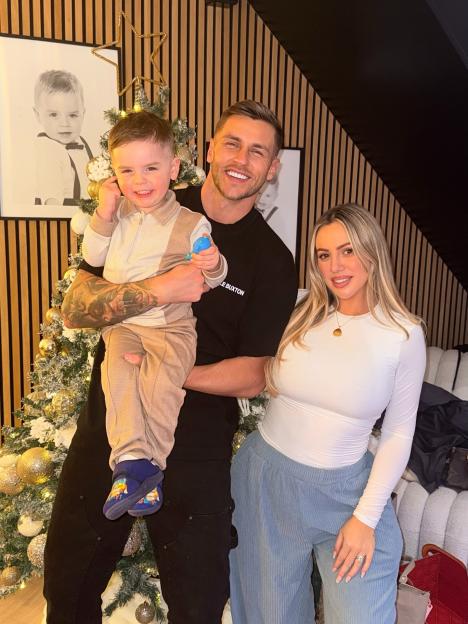 A smiling family of three, parents and their young son, stand in front of a Christmas tree.