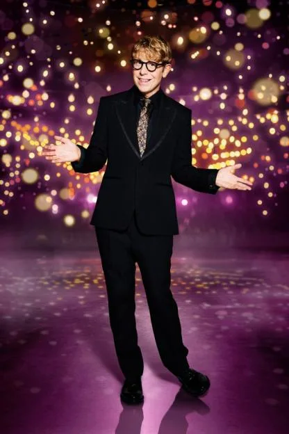Josh Widdicombe in a black suit and patterned tie, smiling with arms outstretched against a backdrop of twinkling lights.