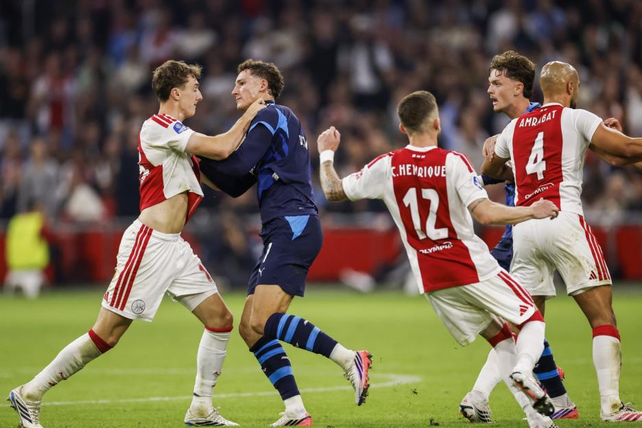 Two AFC Ajax players in red and white uniforms confront an opponent in a blue uniform, while another Ajax player moves to intervene.