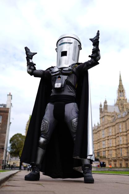 Count Binface, a London mayoral candidate, poses in a full body costume with a bin for a head, black cape, and body armor, with the Houses of Parliament in the background.