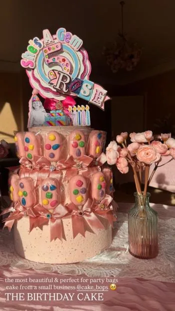 A tiered cake decorated with pink bows and individually wrapped cake pops, topped with a custom sign for Rose's birthday, next to a vase of pink flowers.
