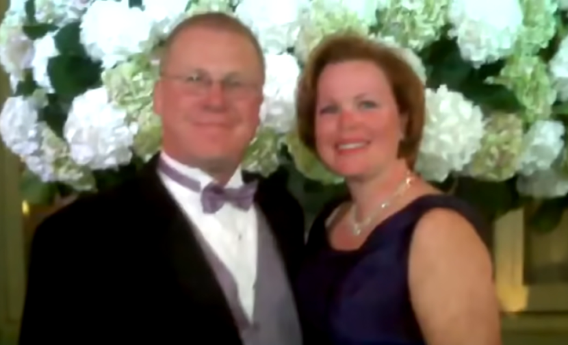 A man and woman smiling in formal attire with white flowers behind them.