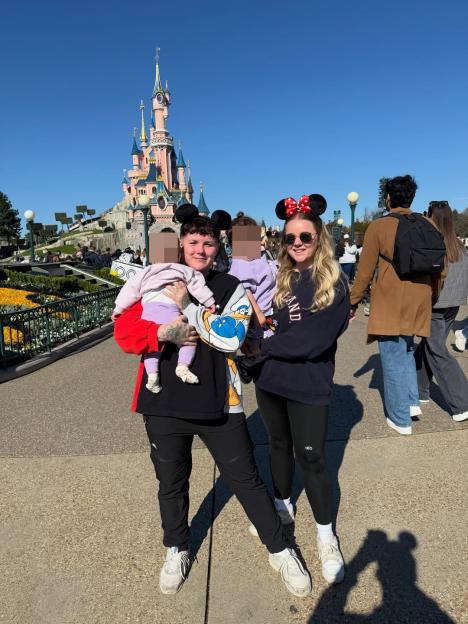 Two women in Mickey and Minnie Mouse ears holding babies in front of a castle.