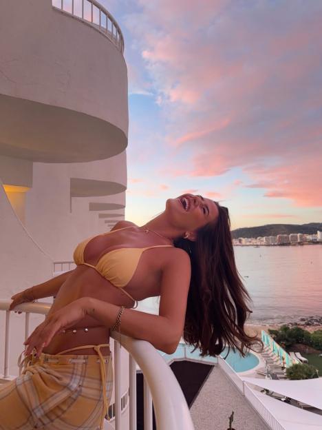 Matilda Draper in a yellow bikini top and plaid pants, laughing on a balcony overlooking the sea at sunset.