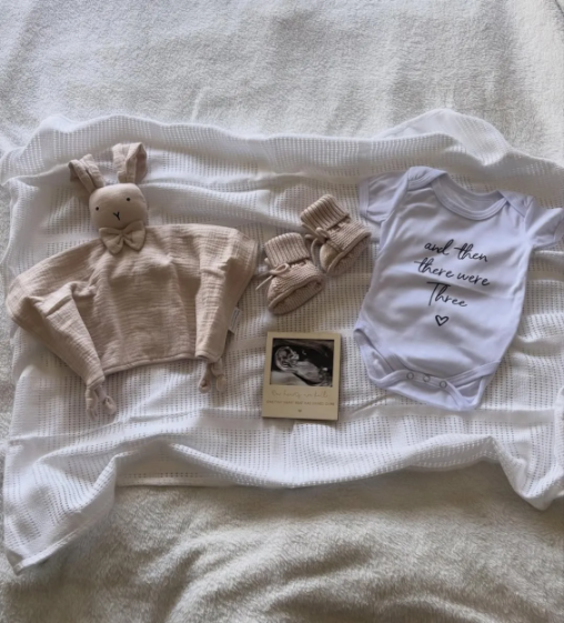 Baby announcement with a onesie that says "And then there were Three", a stuffed rabbit, booties, and an ultrasound photo.