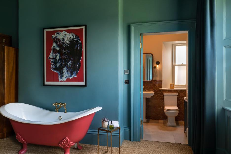 A vibrant red clawfoot bathtub with brass fixtures in a teal room with a large pop-art print of a classical bust, next to a bathroom with a toilet and tiled walls.