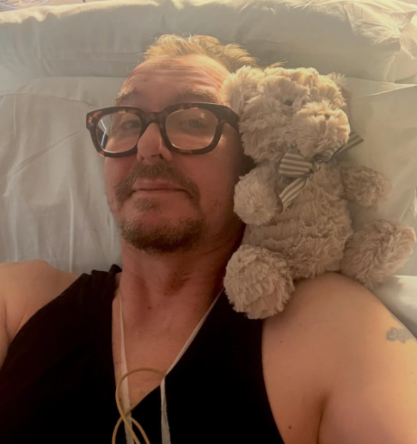 A man in glasses wearing a black top and a teddy bear lie on a light-colored pillow.