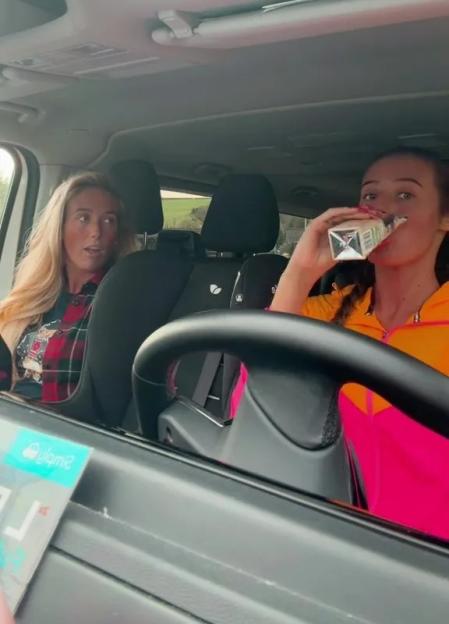 A young woman in the driver's seat drinking from a carton, and an older woman in the passenger seat looking at her.