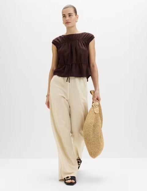 Model wearing a brown top and cream-colored linen trousers with a straw bag.