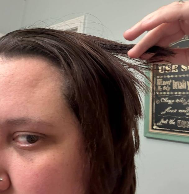 A person's eye and forehead are visible in the foreground, with their hand holding a section of their hair, revealing an uneven and choppy cut.