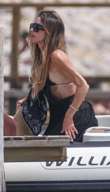 Victoria Beckham in a black bikini top and sheer black dress, wearing sunglasses, standing on a boat.