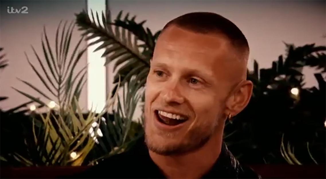 A smiling man with a shaved head and beard, wearing a gold hoop earring, on Love Island All Stars.