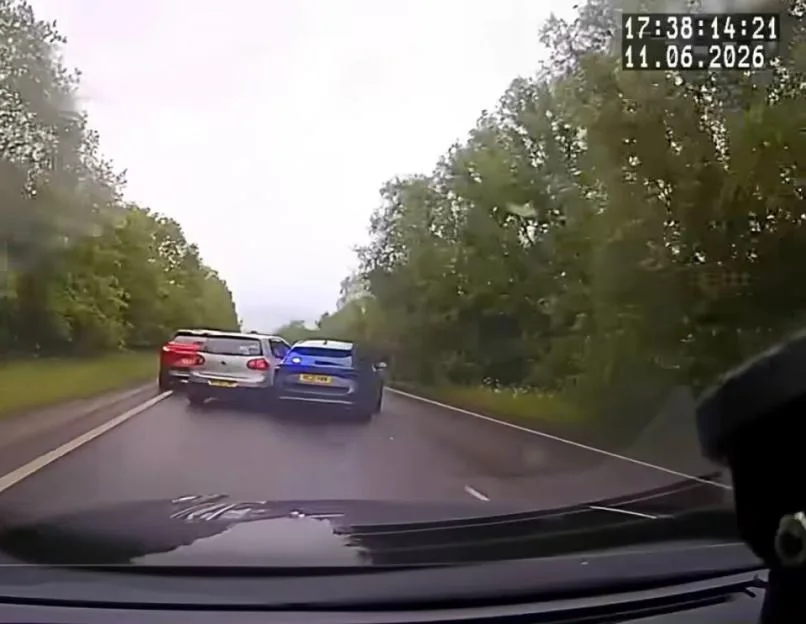 Two cars, one blue and one silver, trapped against a red SUV on a road lined with trees.