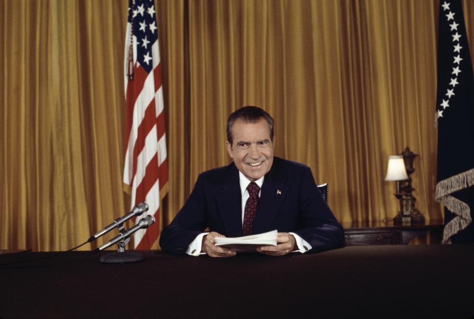 President Nixon smiling at a desk with two microphones in front of him, flanked by American flags.