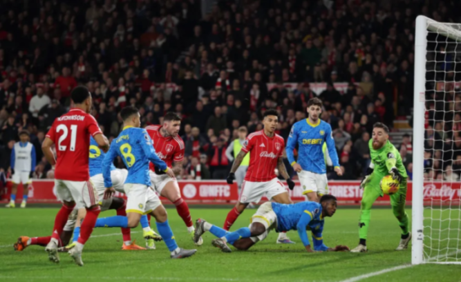 Soccer players in red and blue uniforms jostle for the ball near the goal, one player on the ground, and a goalie in neon green attempting to block.