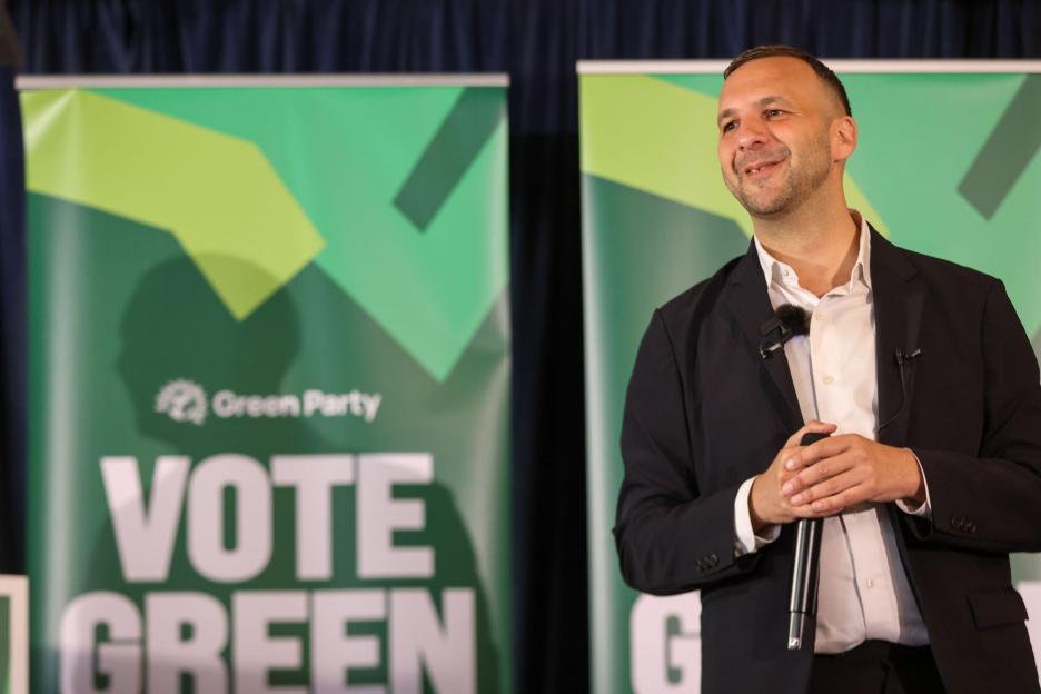 Green Party leader Zack Polanski speaking at an event, with "Vote Green" banners behind him.