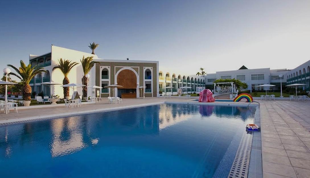 A wide-angle shot of a hotel with a large blue swimming pool in the foreground, palm trees, and outdoor seating with umbrellas.