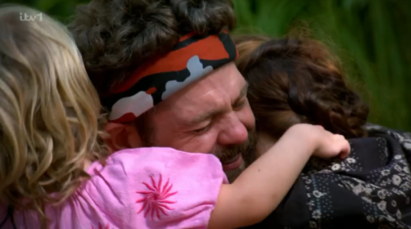 A man with a beard, wearing a camouflage headband, embraces a child in a pink shirt and a woman.