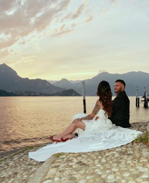 Newlyweds pose by a lake at sunset, with mountains in the background.