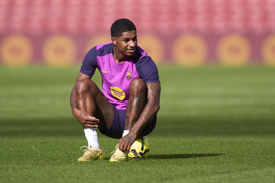 Marcus Rashford in a purple and pink FC Barcelona uniform, crouching on the green grass of a stadium.
