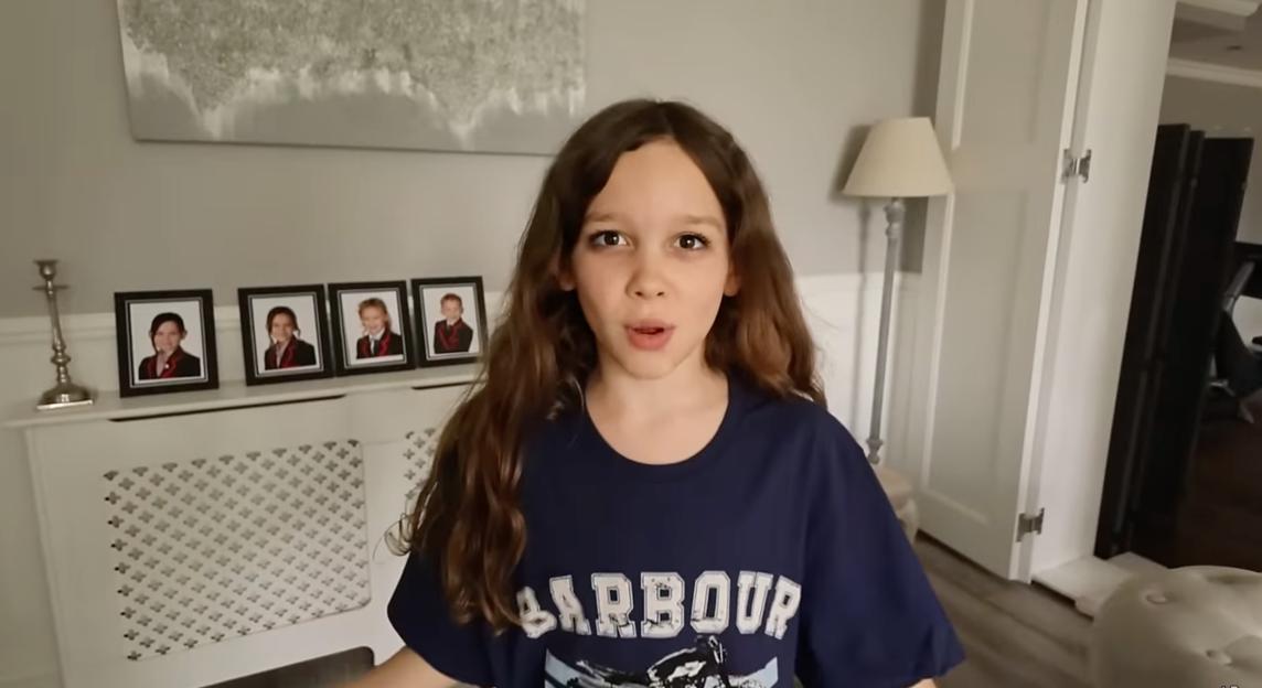 A young girl in a blue shirt with the word "BARBOUR" on it stands in front of a white radiator cover with framed photos on top.