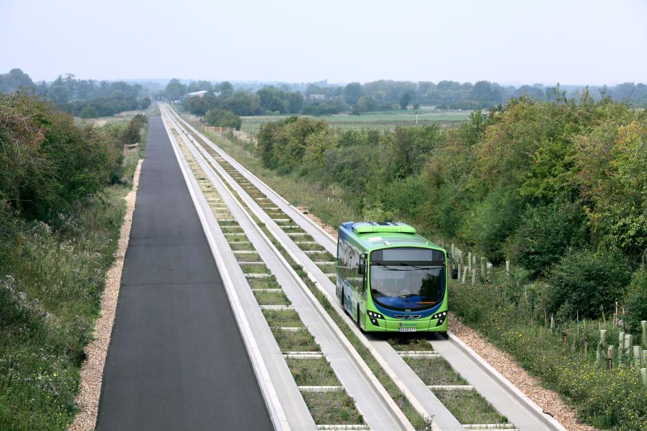 A green and blue bus on a concrete guided busway next to an asphalt path in a rural area.