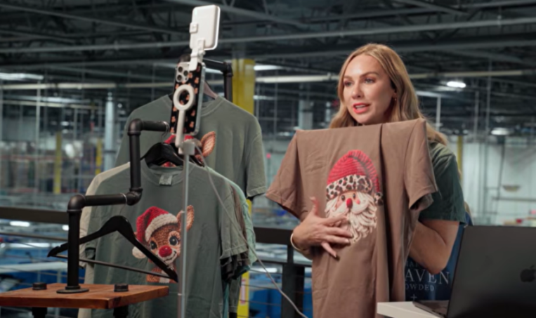 A woman holding a brown t-shirt with a Santa Claus design while filming a video in a warehouse.