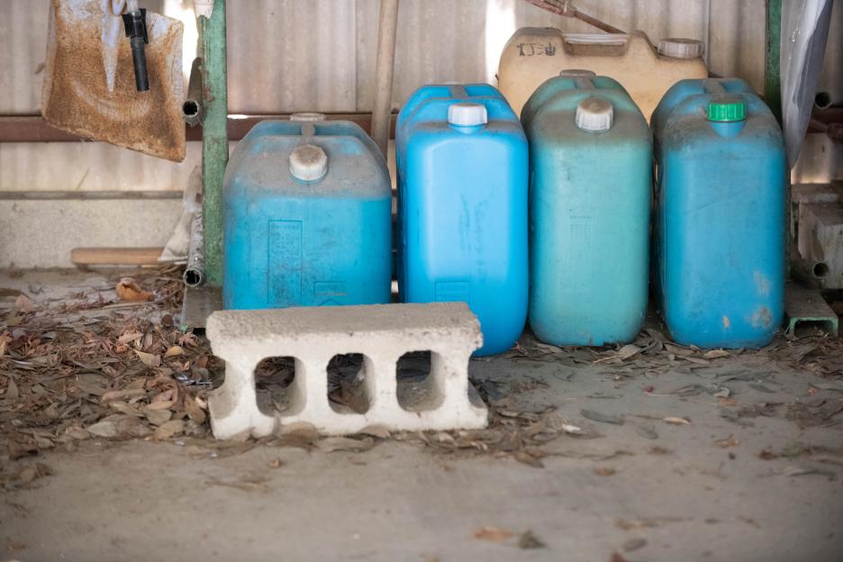 Four blue plastic kerosene tanks and one yellow tank on a dusty concrete floor with fallen leaves, next to a concrete block.