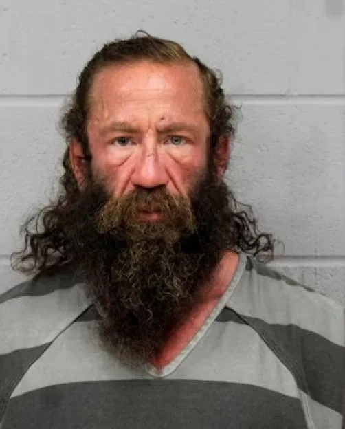 Mugshot of a middle-aged man with long, unkempt brown hair and a thick beard, wearing a striped jail uniform.