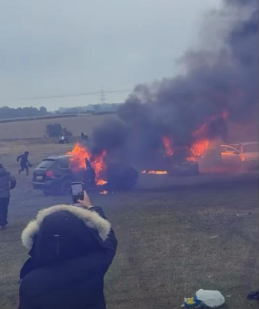 People standing around cars set on fire, emitting dark smoke, in a field.