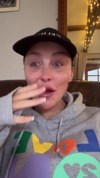Ella Morgan crying and wiping her face, wearing a black hat and a grey hoodie with a colorful "LOVE" design.