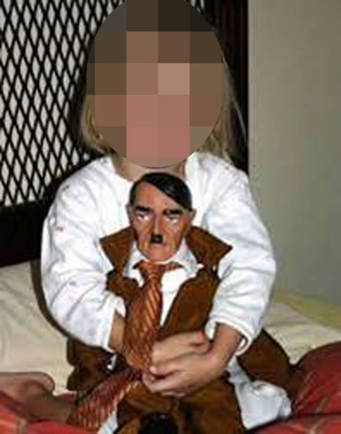 Young child with a pixilated face holds a doll of Adolf Hitler.