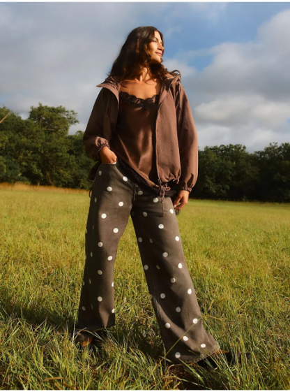 A person wearing a brown jacket, a brown lace camisole, and black wide-leg pants with white polka dots stands in a grassy field.