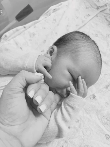 A black and white photo of a newborn baby lying on a blanket, holding onto an adult's hand.