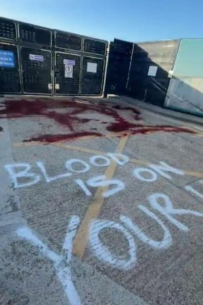 Fake blood splattered on pavement with the words "BLOOD IS ON YOUR" spray-painted, in front of a metal fence.