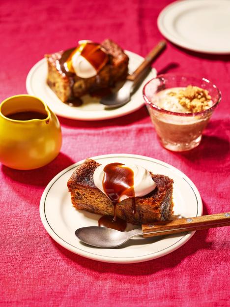 Two slices of sticky toffee pudding with whipped cream and caramel sauce, served on plates with spoons, next to a yellow creamer and a glass dessert.