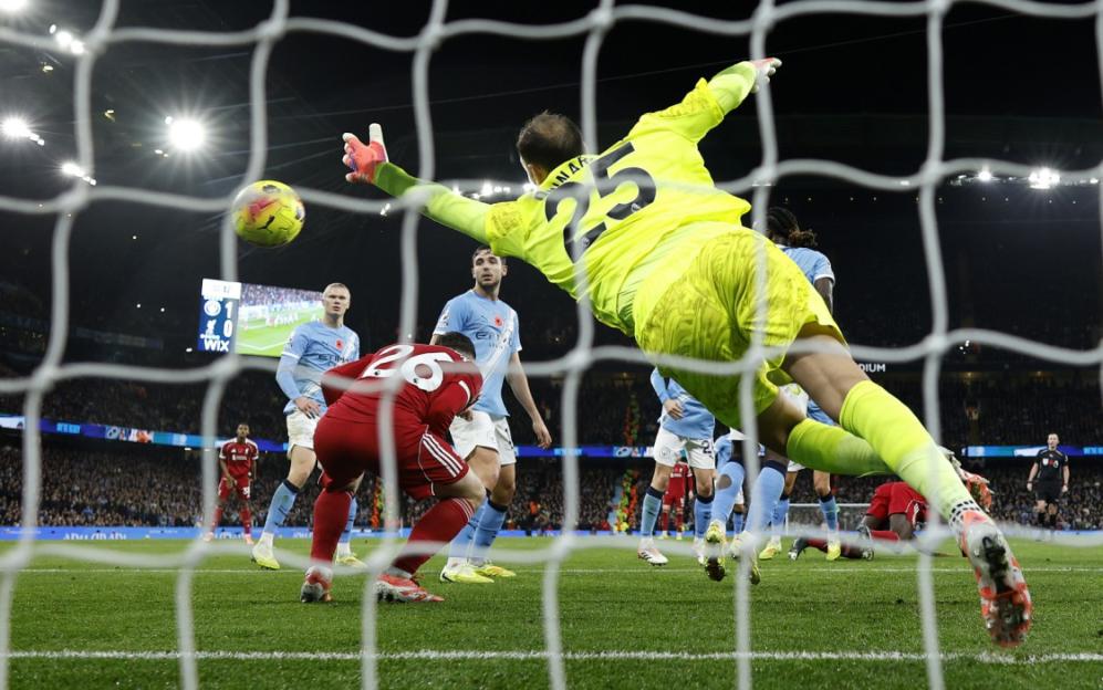 A soccer goalie in a neon yellow uniform dives to block a shot, viewed from behind the net, with other players visible on the field.