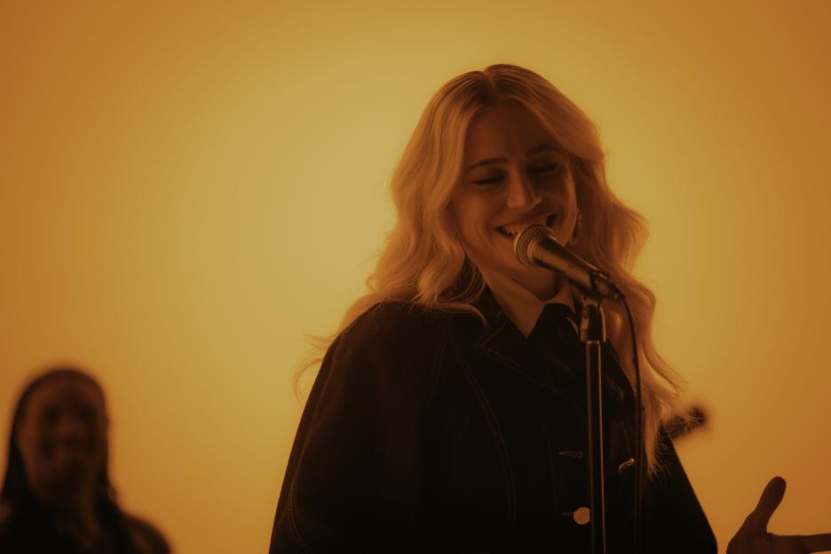 Pixie Lott smiling while singing into a microphone, lit by orange stage lights.