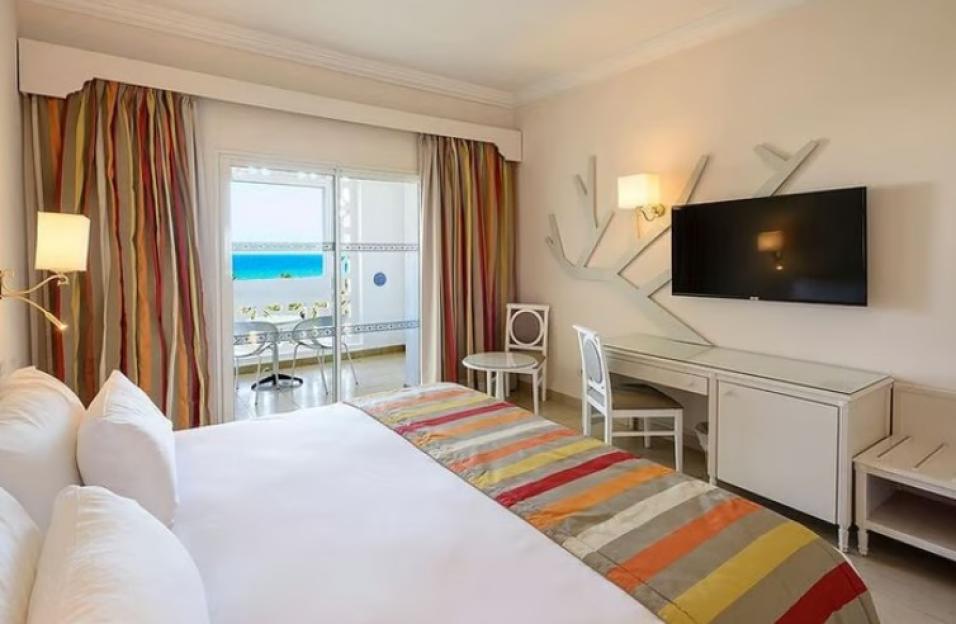 A hotel room with a bed, a desk, a TV, and a balcony overlooking the sea.