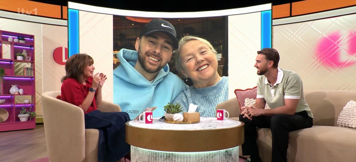 Two people seated on a TV show set, with a large image of a man and an older woman on a screen behind them.