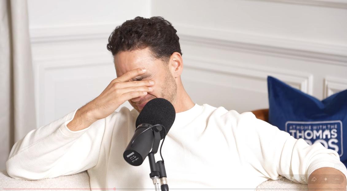 A man in a white ribbed shirt covers his face with his hand, sitting next to a black podcast microphone, with a blue pillow in the background.