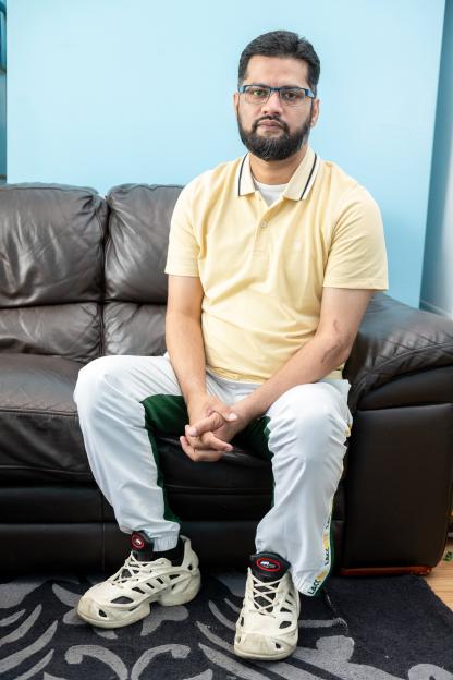Raza Aslam, a dad of four, sits on a brown leather couch in a yellow polo shirt and white and green pants.