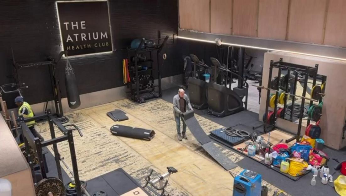 Inside a gym with damaged floors, a man unrolls a mat while cleaning supplies are piled in the foreground.
