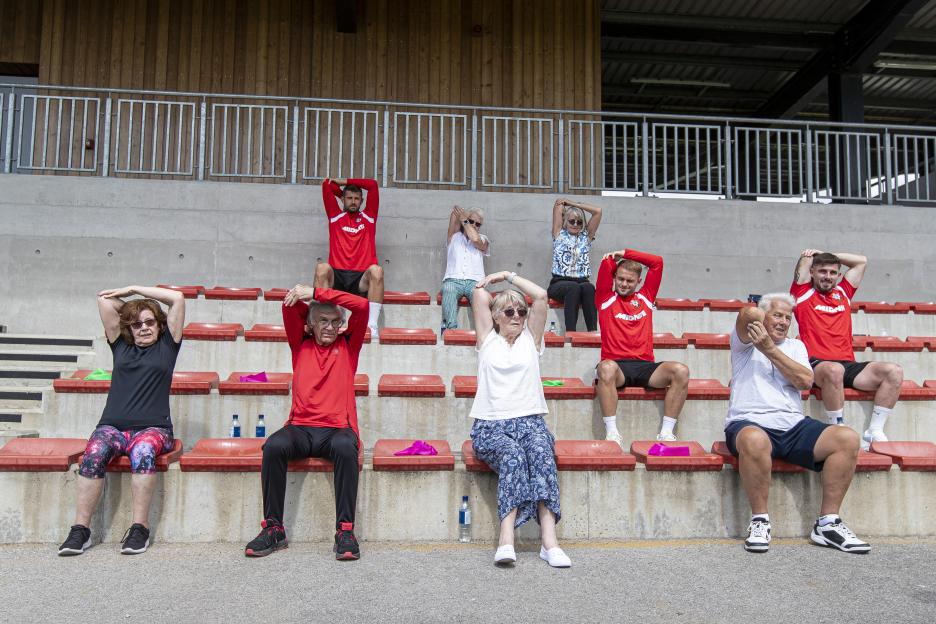 A group of seniors and younger men stretching their arms while sitting on bleachers.
