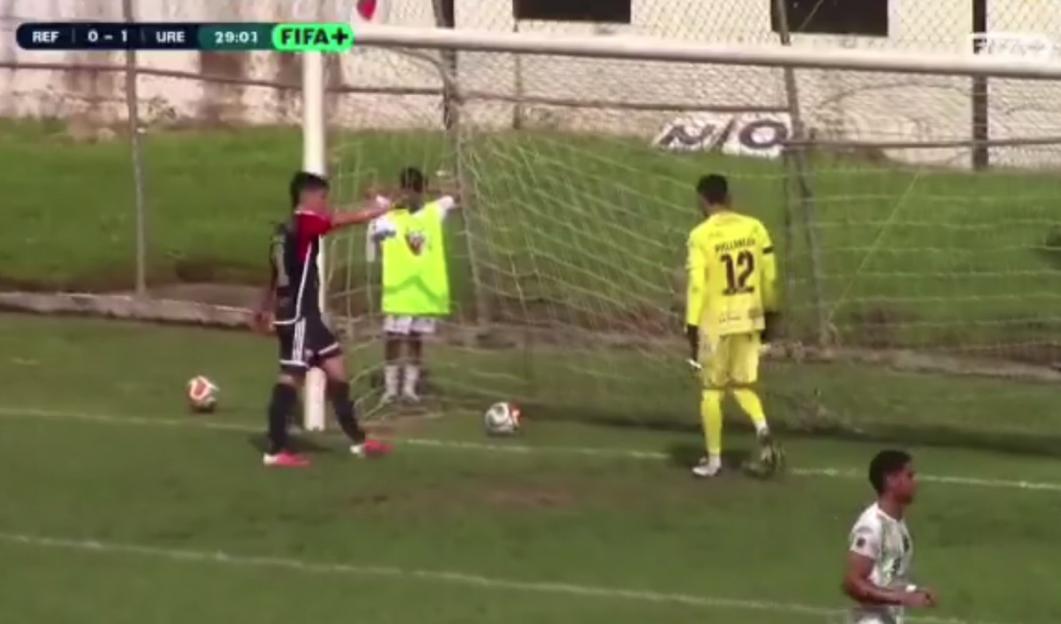 A soccer player in black and red uniform disputes a call with the referee while a ball sits on the goal line.
