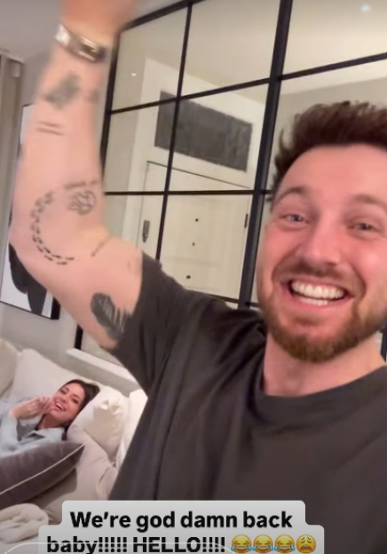 A man in a dark t-shirt with arm tattoos smiling widely, with a woman laughing on a couch behind him.