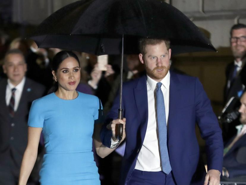 Meghan Markle in a blue dress and Prince Harry in a blue suit walk under an umbrella.