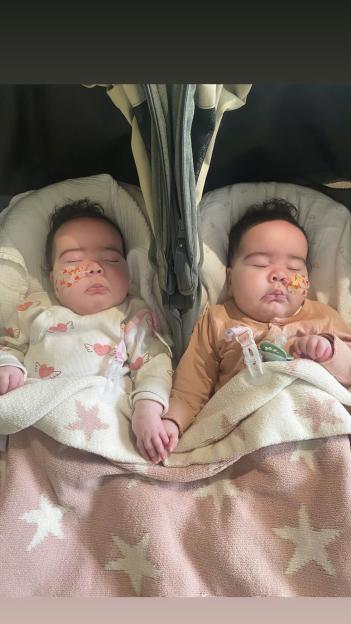 Two twin infants asleep in a stroller, holding hands, with feeding tubes taped to their cheeks.