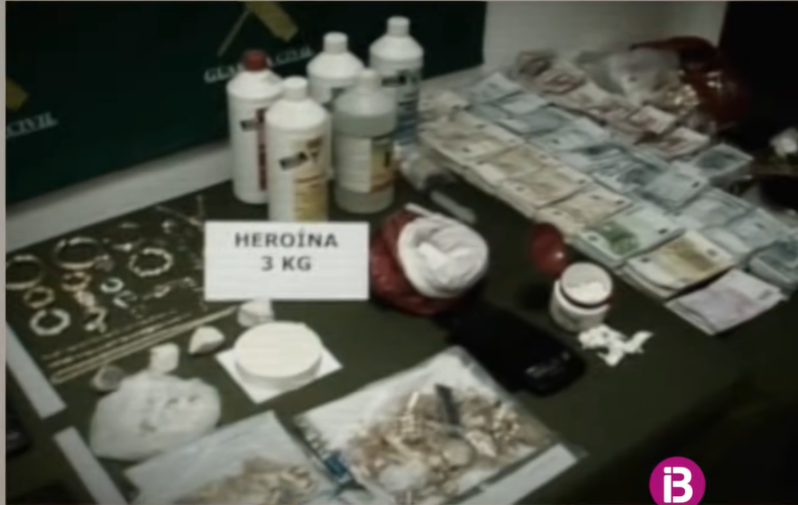A screengrab from a documentary showing criminal proceeds laid out on a table, including bundles of cash, drugs labeled "HEROÍNA 3 KG", white powder, chemicals in bottles, jewelry, and a syringe.